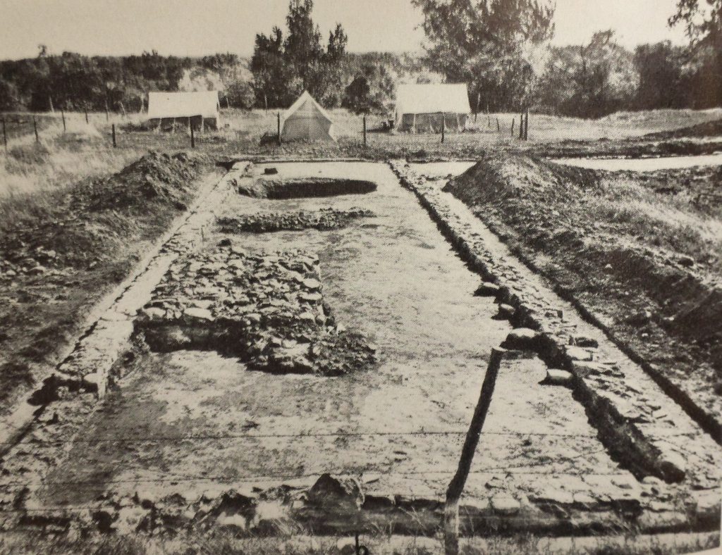 Black-and-white photo of excavation, with excavated area in the foreground and tents in the background.