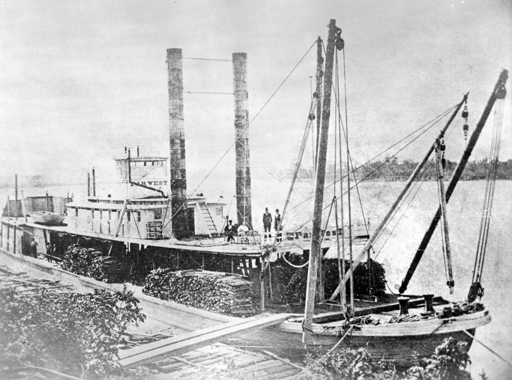An old and thus low-quality black-and-white photo of a docked steamboat with some people on deck and a lot of cargo.