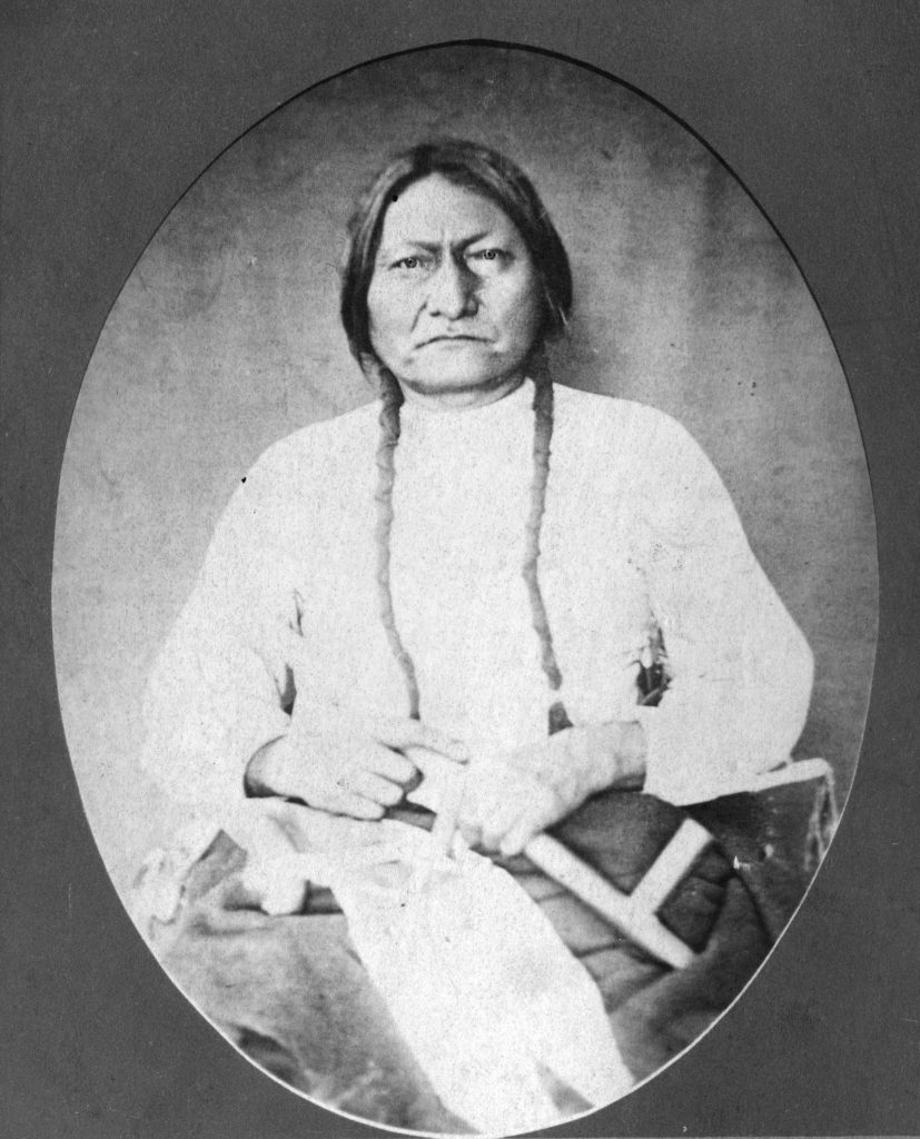 A black-and-white photo in an oval frame. Sitting Bull is seated and shown from approximately the knees up. He is in a white shirt with possibly blankets in his lap and holding a white pipe.