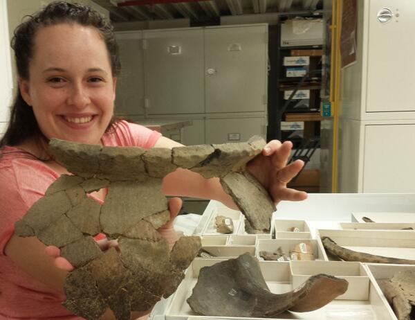 A smiling woman holding up a large, but very partial vessel. Enough sherds have been glued together that it's round shape is clearly visible.