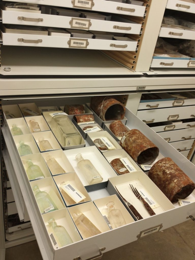 An open drawer in a cabinet with many drawers, surrounded by other cabinets. In the drawer are small cardboard boxes with glass bottles, metal cans, and a couple forks.