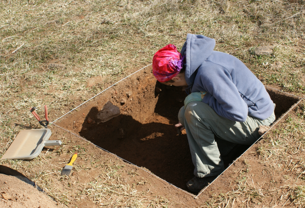 Photo of me in a 1x1 meter excavation unit, looking at the surface I'm working on and thus away from the camera.