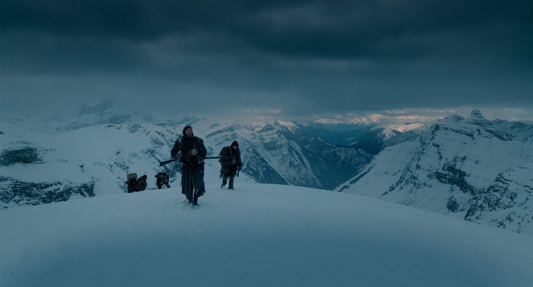 Still from The Revenant. Men traveling through a snowy mountainous landscape under an ominously cloudy sky.