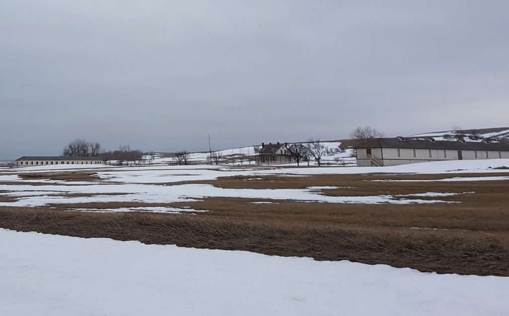 Snowy landscape under a dark grey sky. Long narrow buildings and a large house.
