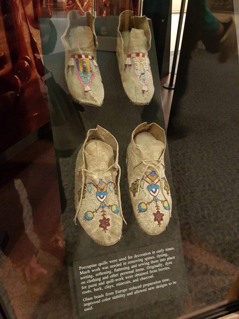This display case contains two pairs of shoes: one decorated with porcupine quills, one with glass beads. Text reads, : Porcupine quills were used for decoration in early times. Much work was needed in removing spines, dyeing, sorting, softening, flattening and sewing them into place on clothing and other personal items. Originally, dyes for paint and quill-work were obtained from berries, roots, bark, clays, minerals, and charcoal. Glass beads from Europe reduced preparation time, improved color stability and allowed new designs to be used."