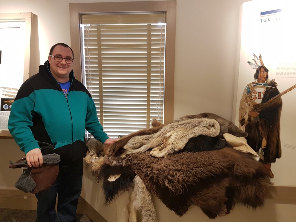 Mat posing with a display that consists of a pile of furs. His left hand is on the furs. He is smiling.
