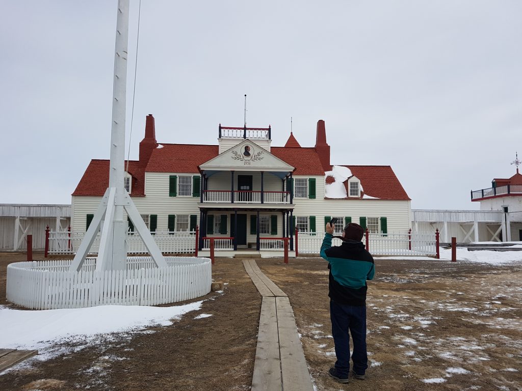 My friend Mat, standing inside the fort, in front of the Bourgeois house, taking photos. It's a two-story building with white siding and bright red roof tiles and chimneys. It has many windows with green shutters and a front patio on both floors, with a triangular roof at the top, There is a picture of the bourgeois on the front of it. There's another exterior space with a railing above that.