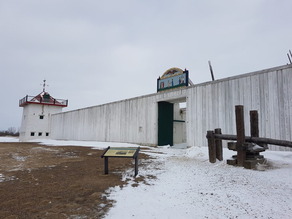 The whitewashed stockade with a bastion and an entrance gate in the center. Above the gate is a painting showing trading partners greeting each other against a pale blue background. In front of the fort is a reconstructed fur press. The landscape is snowy and the sky is grey.