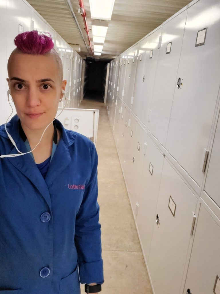 Selfie, standing in a long aisle of museum collection storage cabinets wearing a blue lab coat with my name on the front left.