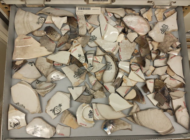 Color photo of ceramic sherds in a cardboard tray in a storage drawer. They are mostly plate bottoms that include maker's marks and they are positioned so that the marks are facing up.