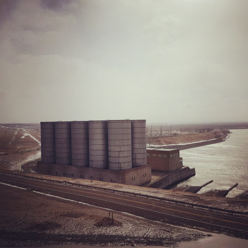 The power house at Garrison Dam, a blocky construction with ten cylindrical towers closely spaced together, in low winter light under a grey sky.