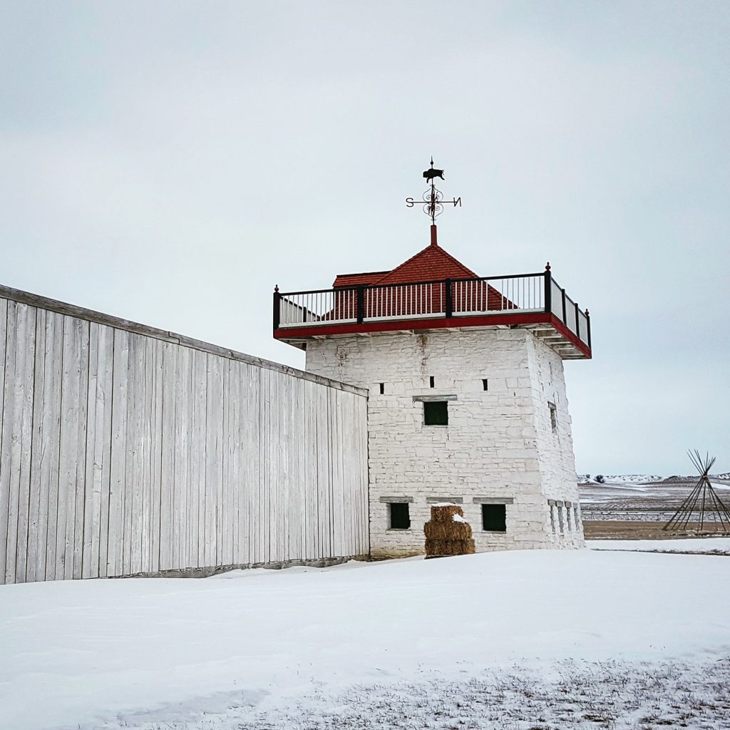 One of the whitewashed brick bastions at Fort Union. It has a pointy red tile roof with a large weathervane. The weathervane has a bison on it. The skeleton of a teepee stands off to the side in the snowy landscape. The sky is grey.