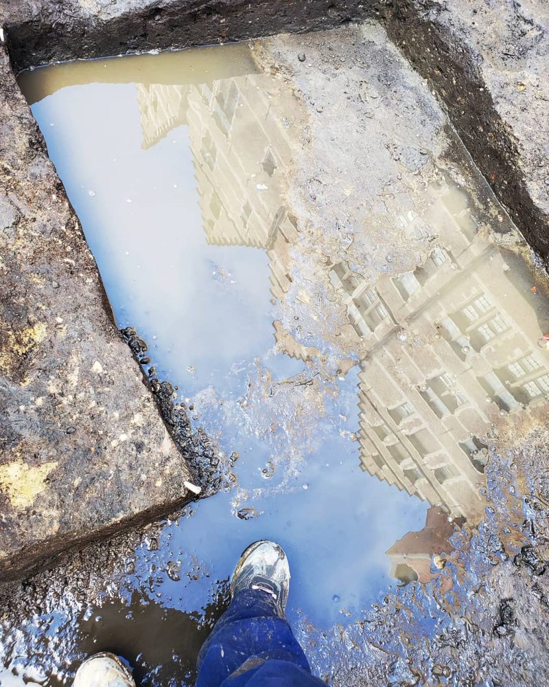 Looking down at my feet, in steel-toed boots, in a very muddy excavation unit. It has standing water in it. Sixteenth-century building facades and partially blue sky are reflected in the water.