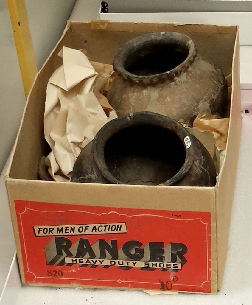 Two mended ceramic pots stored in a shoe box. A label on the end of the box facing the camera reads "For men of action. Ranger heavy duty shoes" on a bright orange background.