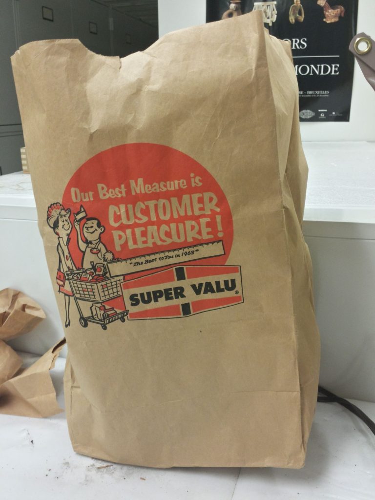A brown paper grocery bag from a store called Super Valu. It has the store name next to an image of a person shopping and the words "Our Best Measure is CUSTOMER PLEASURE" in large print on a bright orange background. Below that text is an image of a ruler with the words "'The Best to You in 1963'".