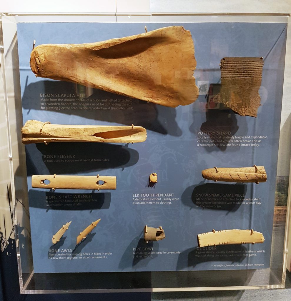 Straight-on view of bone and ceramic artifacts attached to a vertical display with text explanations. They include a bison scapula hoe, a bone flesher and shaft wrench, bone awls, a bone pendant and game piece, and ice glider, a pipe bowl, an elk tooth pendant, and a rim sherd.