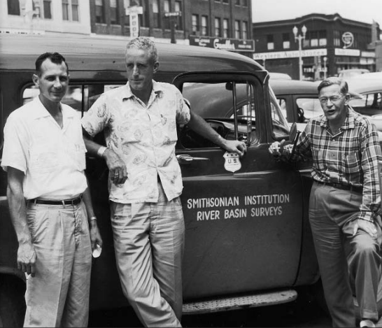 Black-and-white photo of three men leaning against a dark-colored truck with the words "Smithsonian Institution River Basin Surveys" on it.