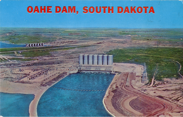 Color postcard. View of Oahe dam with power house in the center of the image. Some areas on the edge o f the lake look like they are still under construction. Text reads, "Oahe Dam, South Dakota" in big red block letters across the blue sky in the top quarter of the image.