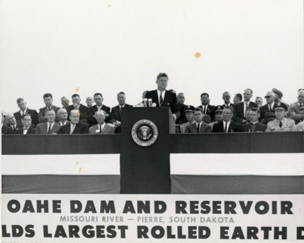 Black-and-white photo of President John F. Kennedy at a lectern, with several rows of people seated and standing behind him. A partial banner is visible on the front of the podium they are all on, which reads "Oahe Dam and Reservoir. Missouri River - Pierre, South Dakota." Below that the text is cut off on both sides butit presumably reads, "World's largest rolled earth dam".