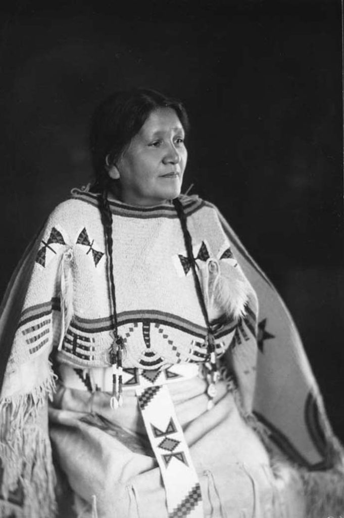Black-and-white portrait of Josephine Gates Kelly, seated. Her hair is in two braids and she is wearing light-colored embroidered/beaded clothing.