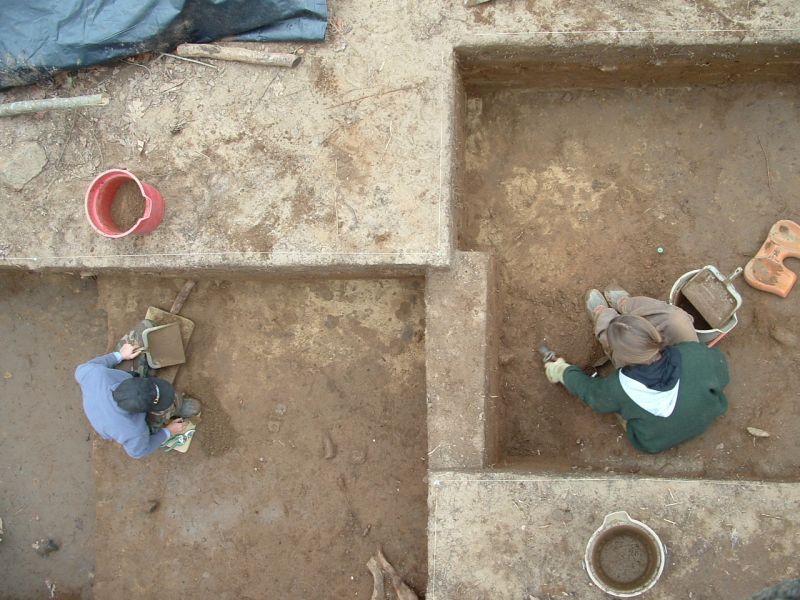 A top down view of the author and a colleague working in adjoining test units, offset by a half unit (because of the shape of the riverbank, not in the photo). White string follows the outline of the test units. The units are excavated to the same depth (40 cm or so) but a small baulk is left in place between the two.