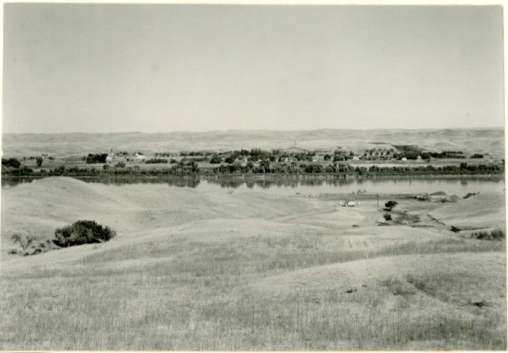 Black-and-white landscape photo, showing undulating grasslands in the foreground, the Missouri river with some buildings on both river banks.