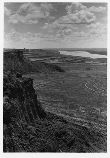 Black-and-white landscape photo, showing river bluffs with digging marks and loose soil full of big tire imprints. The river is in the background.