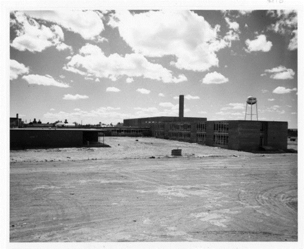 Black-and-white photo of a complex of cuboid two-story buildings with a chimney on one. A watertower is also visible behind the buildings. Dusty open space in the foreground.