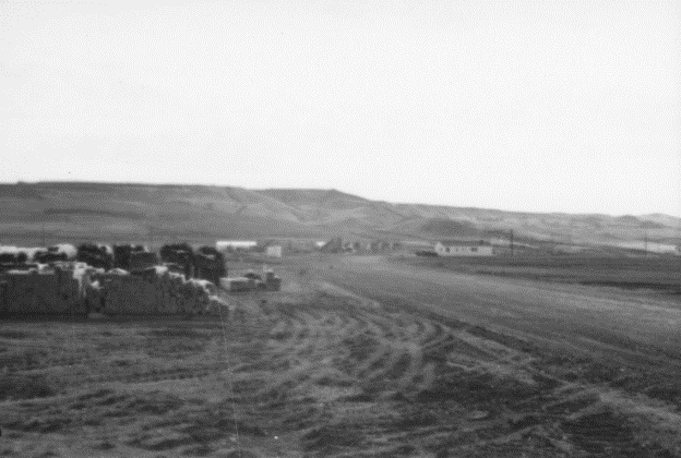 Black-and-white photo of construction material in a large dusty area marked by tire tracks. Low hills in the background.