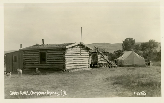 Black-and-white photo of a log cabin with several lean-to structures and a tent behind it. A couple of dogs stand in the foreground. The words "Sioux Home, Cheyenne Agency, S.D." are written in white ink, along with what appears to be a signature.