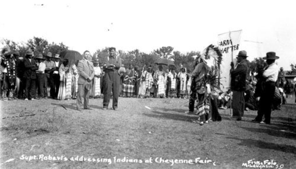 Black-and0white photo of a large group of people standing in a field. Some are in suits, some in regalia.