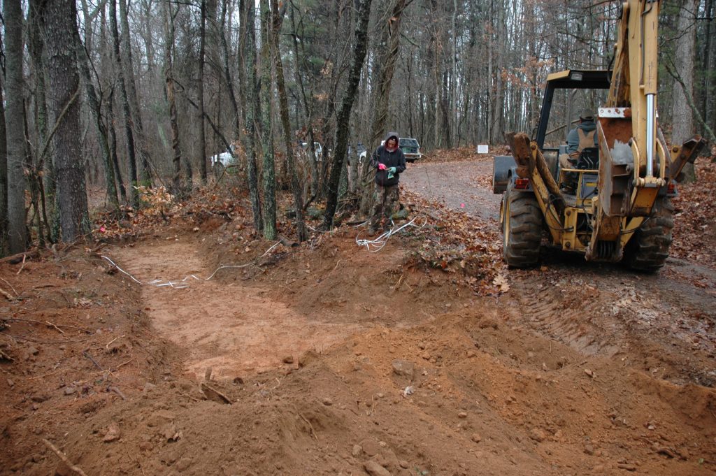 A forest scene with a dirt/gravel road to the right and a test trench dug by a backhoe to the left. The backhoe is driving away onto the road while the author stands by the trench rolling up string and a long tape measure used to measure across long distances. Vehicles are partially visible parked in the background.