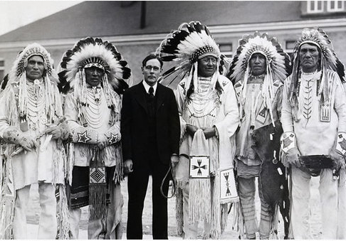 Six people standing side by side. John Collier is in the middle in a black suit. The other five people are chiefs in full regalia. They are wearing light colored clothing and feather headdresses.