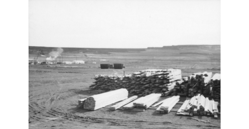 Black-and-white photo of s tacks of lumber lying in a dusty flat area full of  tire tracks. Low buildings, smoke, and other signs of activity in the background.