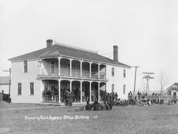 Black and white photo of two-story white building with porches on both floors. Many people are standing on the lower one. Others are seated on the grass in front of the building.