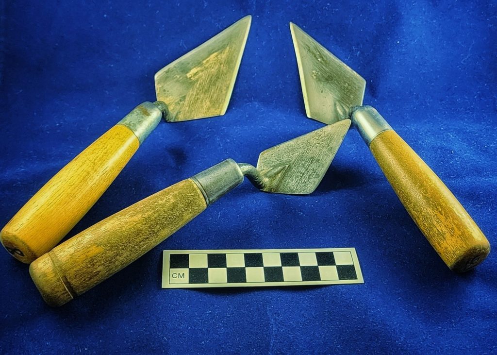 Three slightly dirty trowels on a blue background with a photo scale.