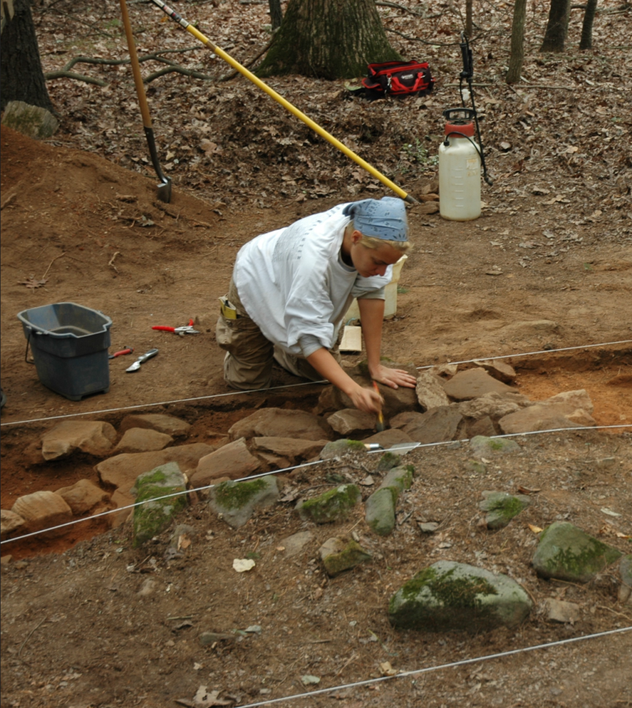 The author, kneeling over a test unit (the same one as pictured before with the chimney rocks in the center), brushing soil off rocks. An unexcavated part of the test unit is in the foreground with rocks sticking up out of the surface.