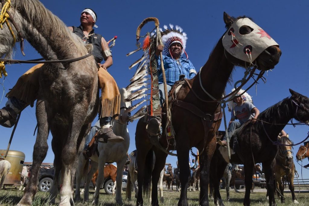 Low-angle photo of men on horseback against a deep blue sky. In the center is Harold Frazier in a blue plaid shirt and a large feather headdress.