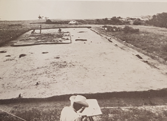 Black-and-white photo of the excavation area with people working in it. Topsoil has been stripped off a large rectangular area.
